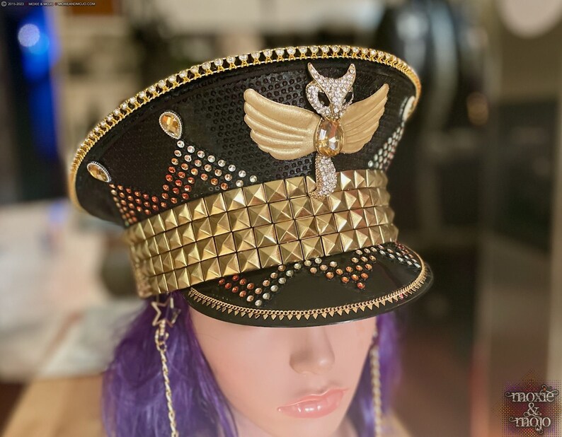 May include: A black and gold sequined hat with a gold studded band and a winged angel design. The hat is decorated with rhinestones and has a gold chain attached to the side.