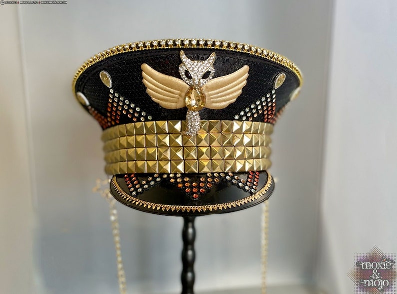 May include: A black and gold embellished hat with a gold studded band, a gold winged owl with a crystal design, and a chain hanging from the brim. The hat is on a black stand.