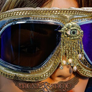 May include: Gold-toned goggles featuring dark purple lenses, a metallic frame, and a gold eye-shaped embellishment with dangling chains and small white pearls. The goggles are trimmed with gold chain detailing and include a tan elastic strap.