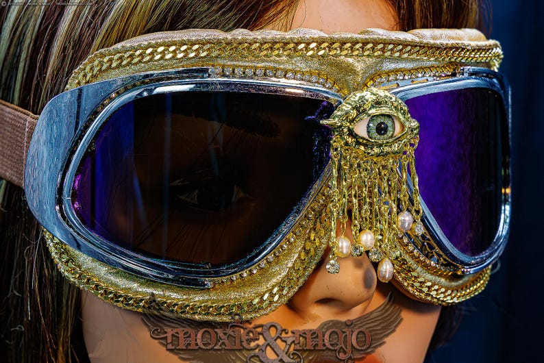 May include: Gold-toned goggles featuring dark purple lenses, a metallic frame, and a gold eye-shaped embellishment with dangling chains and small white pearls. The goggles are trimmed with gold chain detailing and include a tan elastic strap.