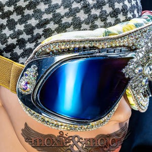 Puede incluir: Gafas protectoras con lentes reflectantes azul oscuro, marco plateado y correa el&aacute;stica dorada. El marco est&aacute; decorado con pedrer&iacute;a transparente e iridiscente y un gran adorno de estrella de cristal central. El texto dice moxie & mojo.