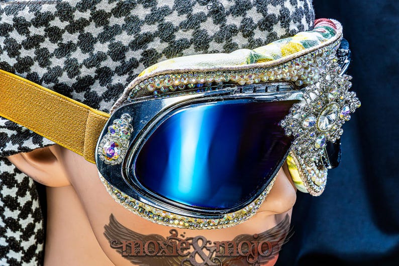 Puede incluir: Gafas protectoras con lentes reflectantes azul oscuro, marco plateado y correa el&aacute;stica dorada. El marco est&aacute; decorado con pedrer&iacute;a transparente e iridiscente y un gran adorno de estrella de cristal central. El texto dice moxie & mojo.