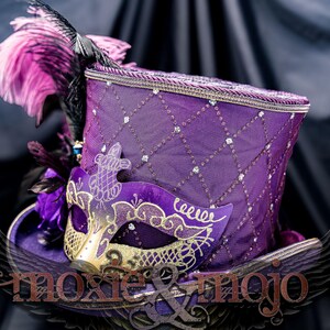 May include: A purple top hat featuring a diamond-patterned mesh overlay with embedded crystals, adorned with pink and black feathers and a purple and gold masquerade mask. The text moxie & mojo is visible across the lower portion of the hat.