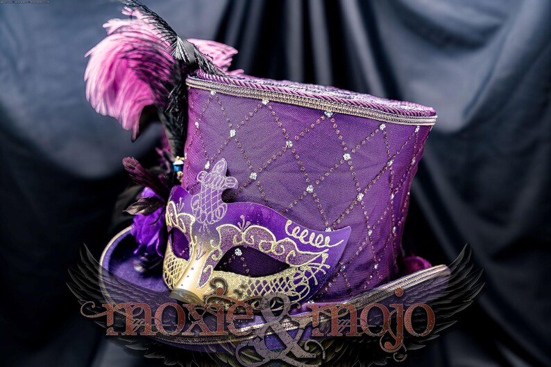 May include: A purple top hat featuring a diamond-patterned mesh overlay with embedded crystals, adorned with pink and black feathers and a purple and gold masquerade mask. The text moxie & mojo is visible across the lower portion of the hat.