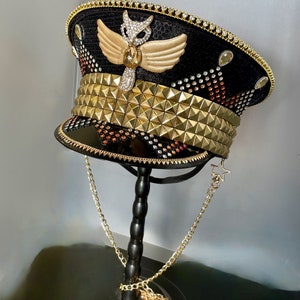 May include: A black and gold sequined hat with a gold owl design and a gold chain. The hat is decorated with rhinestones and has a gold band with pyramid-shaped studs.