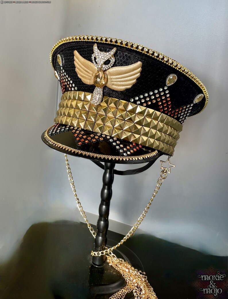 May include: A black and gold sequined hat with a gold owl design and a gold chain. The hat is decorated with rhinestones and has a gold band with pyramid-shaped studs.
