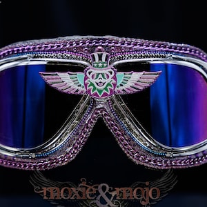 Puede incluir: Gafas protectoras moradas con montura cromada y lentes azules reflectantes. El puente central muestra un emblema de oso alado con sombrero de copa. La montura est&aacute; adornada con cadenas moradas y peque&ntilde;os remaches. El texto moxie & mojo es visible.