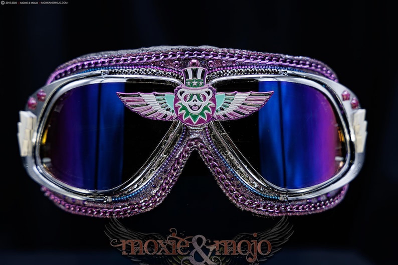 Puede incluir: Gafas protectoras moradas con montura cromada y lentes azules reflectantes. El puente central muestra un emblema de oso alado con sombrero de copa. La montura est&aacute; adornada con cadenas moradas y peque&ntilde;os remaches. El texto moxie & mojo es visible.