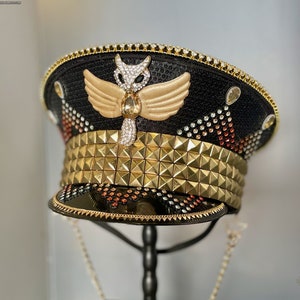 May include: A black and gold embellished hat with a gold studded band and a gold owl with wings on the top. The hat is on a black stand and has a gold chain attached to it. The text "moxie & mojo" is visible in the bottom right corner of the image.