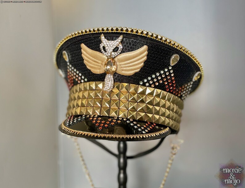 May include: A black and gold embellished hat with a gold studded band and a gold owl with wings on the top. The hat is on a black stand and has a gold chain attached to it. The text "moxie & mojo" is visible in the bottom right corner of the image.