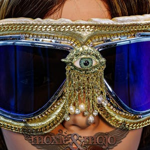 May include: Goggles with dark blue reflective lenses, a gold-toned frame, and a central eye emblem. The frame is decorated with gold chains and rhinestones. A gold-toned fringe with small white pearls and clear crystals hangs from the eye ornament.