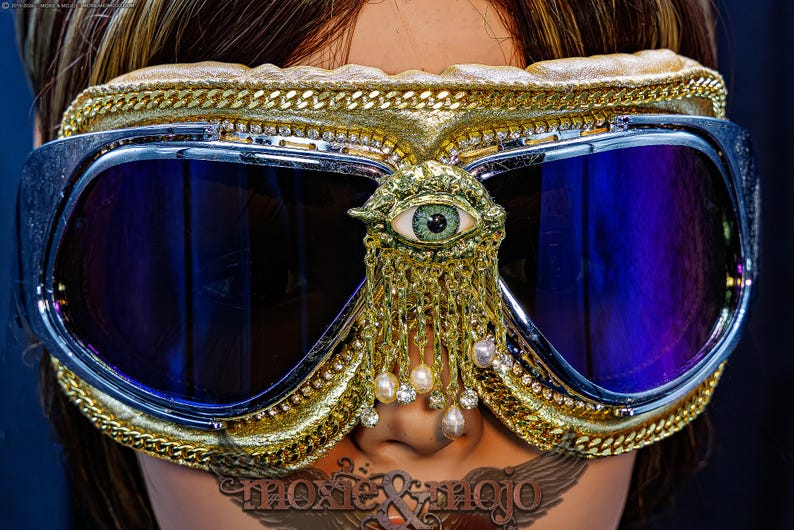 May include: Goggles with dark blue reflective lenses, a gold-toned frame, and a central eye emblem. The frame is decorated with gold chains and rhinestones. A gold-toned fringe with small white pearls and clear crystals hangs from the eye ornament.