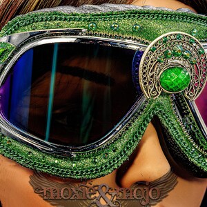 Pu&ograve; includere: Occhiali protettivi verdi con lenti scure e montatura argentata. L'esterno &egrave; decorato con glitter verdi, piccoli strass verdi e un vistoso ornamento in filigrana color argento antico con una grande pietra preziosa ovale di colore verde.