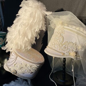 May include: Two white marching band hats with elaborate white beading and trim. One hat has a white feather plume and the other has a white tulle veil.
