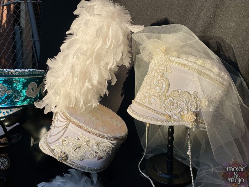 May include: Two white marching band hats with elaborate white beading and trim. One hat has a white feather plume and the other has a white tulle veil.