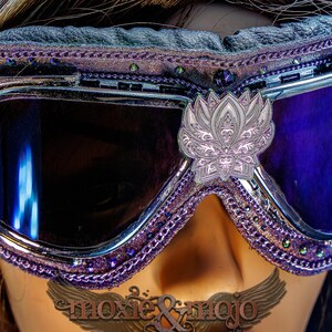 Puede incluir: Gafas protectoras de color morado y plateado con lentes oscuras y un emblema central de flor de loto plateada. El marco est&aacute; decorado con bordes morados, peque&ntilde;os cristales incrustados y un acolchado negro en el borde superior.