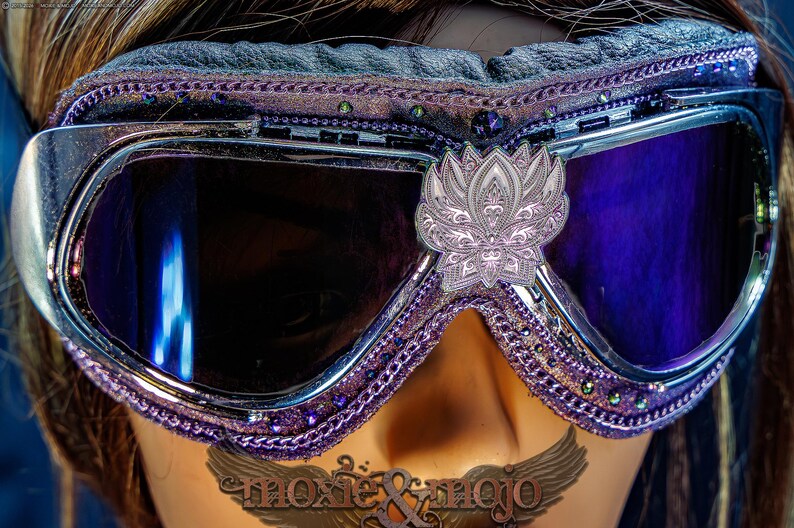 Puede incluir: Gafas protectoras de color morado y plateado con lentes oscuras y un emblema central de flor de loto plateada. El marco est&aacute; decorado con bordes morados, peque&ntilde;os cristales incrustados y un acolchado negro en el borde superior.