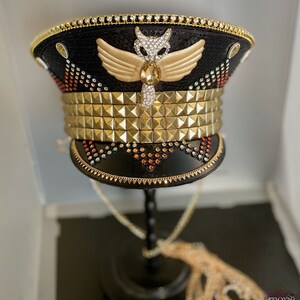 May include: A black and gold embellished hat with a gold winged owl design and a row of gold pyramid studs. The hat is on a black stand with a gold chain.