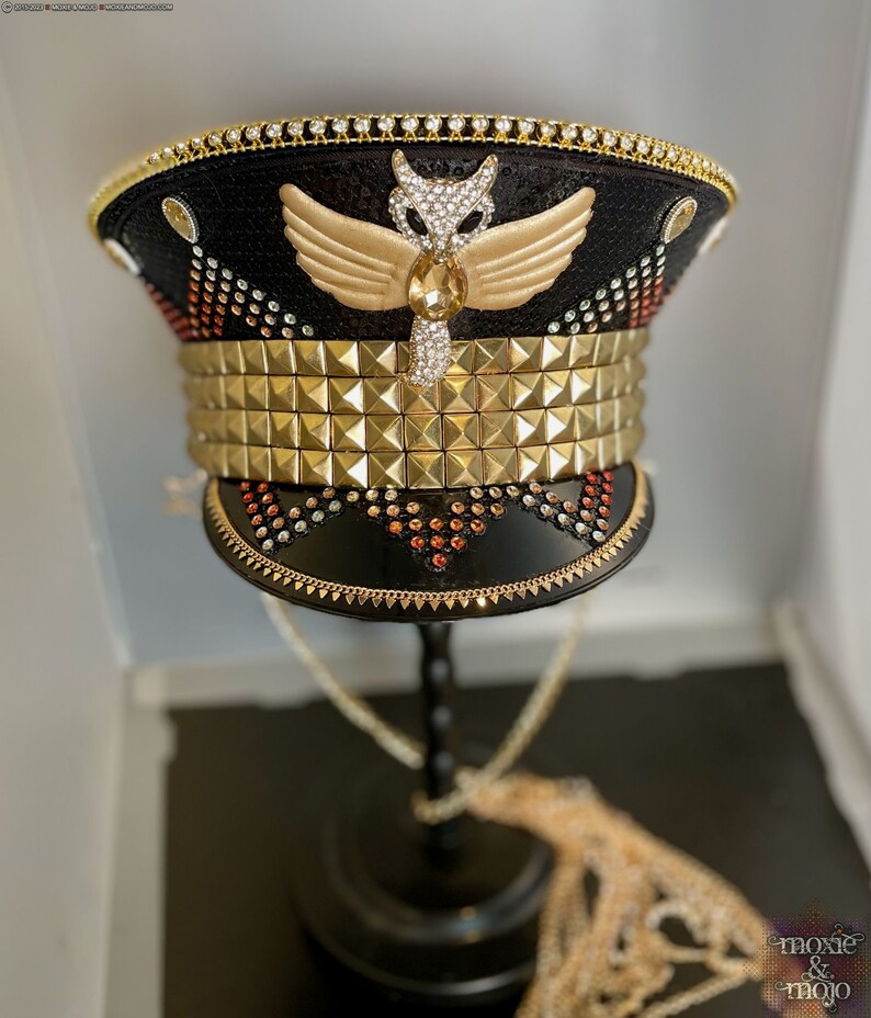 May include: A black and gold embellished hat with a gold winged owl design and a row of gold pyramid studs. The hat is on a black stand with a gold chain.