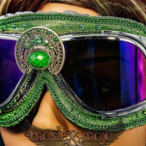 Pu&ograve; includere: Occhiali protettivi verdi con montatura in metallo argentato e lenti color viola scuro. La montatura &egrave; decorata con glitter verdi, piccoli strass verdi e un emblema centrale in filigrana color oro con una grande pietra verde ovale.
