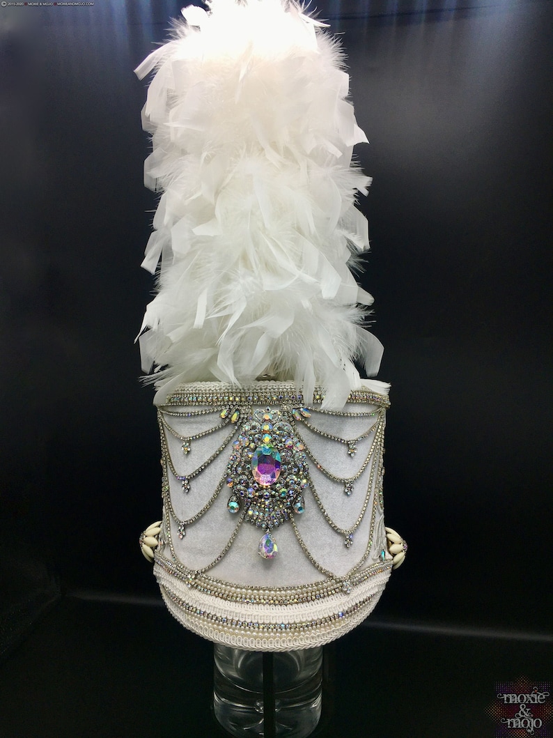May include: A white feather plume sits atop a white hat with a silver trim and a large iridescent jewel in the center. The hat is decorated with silver chains and beads.