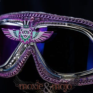 Puede incluir: Gafas protectoras moradas con lentes oscuras, marco cromado y un emblema central de un oso alado con sombrero de copa. Los bordes est&aacute;n decorados con cadenas moradas y peque&ntilde;as estrellas, con un emblema de rayo en el lateral.