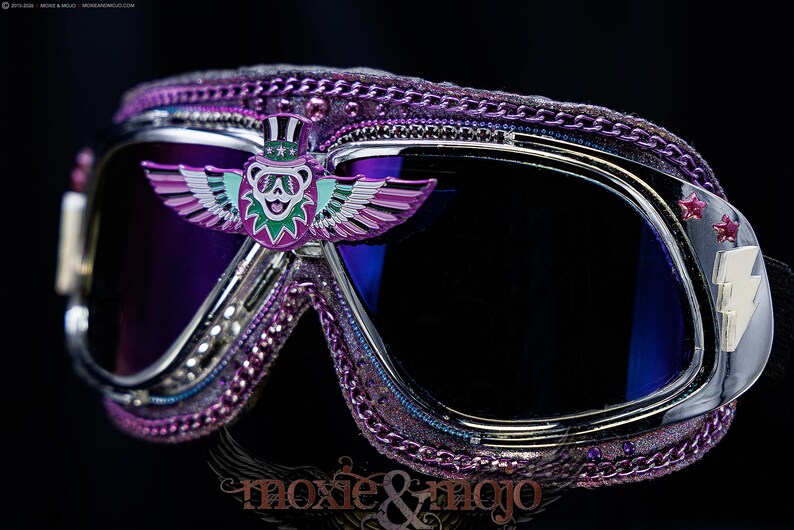 Puede incluir: Gafas protectoras moradas con lentes oscuras, marco cromado y un emblema central de un oso alado con sombrero de copa. Los bordes est&aacute;n decorados con cadenas moradas y peque&ntilde;as estrellas, con un emblema de rayo en el lateral.
