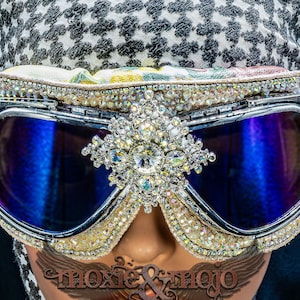 Puede incluir: Gafas protectoras con lentes reflectantes azul oscuro, montura plateada y un adorno central de cristal. Los bordes est&aacute;n incrustados con numerosos peque&ntilde;os diamantes de imitaci&oacute;n transparentes y la parte superior tiene un borde de tela floral.