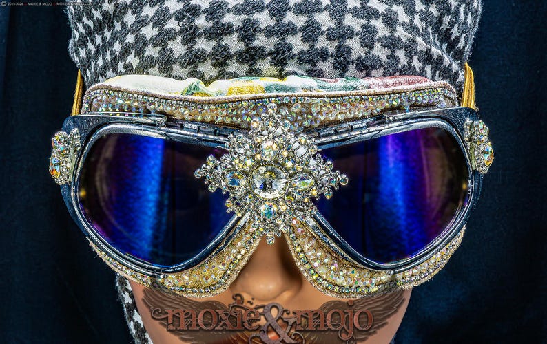 Puede incluir: Gafas protectoras con lentes reflectantes azul oscuro, montura plateada y un adorno central de cristal. Los bordes est&aacute;n incrustados con numerosos peque&ntilde;os diamantes de imitaci&oacute;n transparentes y la parte superior tiene un borde de tela floral.