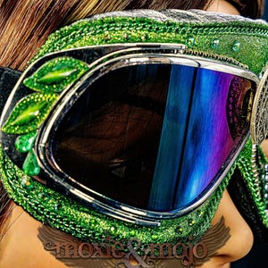 Pu&ograve; includere: Occhiali protettivi verdi con montatura glitterata, decorazioni a forma di foglia e un dettaglio laterale in filigrana con una pietra verde centrale. Le lenti sono scure con riflessi blu e una fascia elastica nera.