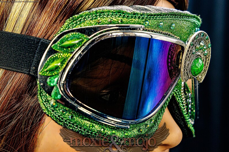 Pu&ograve; includere: Occhiali protettivi verdi con montatura glitterata, decorazioni a forma di foglia e un dettaglio laterale in filigrana con una pietra verde centrale. Le lenti sono scure con riflessi blu e una fascia elastica nera.