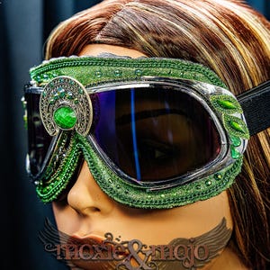 Pu&ograve; includere: Occhiali protettivi verdi con montatura argentata, lenti scure e cinturino elastico. La montatura &egrave; decorata con tessuto verde, intricati filigrani argentati e pietre preziose verdi. Il testo moxie & mojo &egrave; visibile nella parte inferiore.
