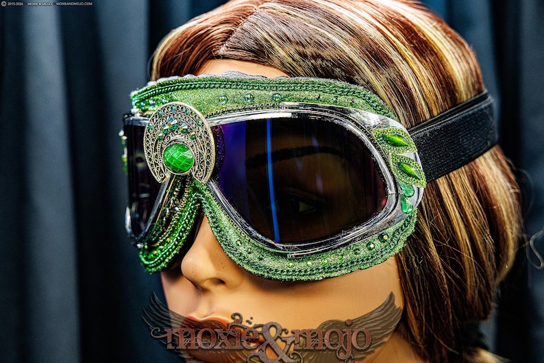 Pu&ograve; includere: Occhiali protettivi verdi con montatura argentata, lenti scure e cinturino elastico. La montatura &egrave; decorata con tessuto verde, intricati filigrani argentati e pietre preziose verdi. Il testo moxie & mojo &egrave; visibile nella parte inferiore.