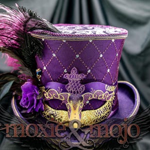 May include: A purple top hat featuring a diamond-quilted pattern with embedded crystals, adorned with a gold and purple masquerade mask, a purple fabric rose, and large black and pink feathers. The text moxie & mojo is visible at the bottom.