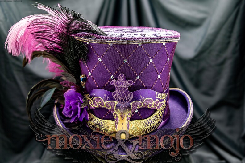 May include: A purple top hat featuring a diamond-quilted pattern with embedded crystals, adorned with a gold and purple masquerade mask, a purple fabric rose, and large black and pink feathers. The text moxie & mojo is visible at the bottom.