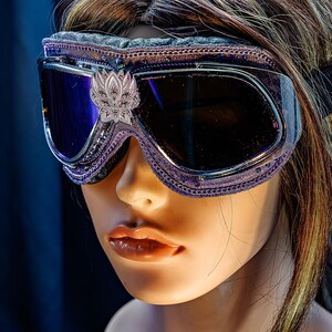 Puede incluir: Gafas protectoras de color morado con lentes oscuras y un emblema plateado en forma de loto centrado en el puente. El marco presenta un borde morado texturizado, detalles met&aacute;licos intrincados y peque&ntilde;os cristales incrustados.