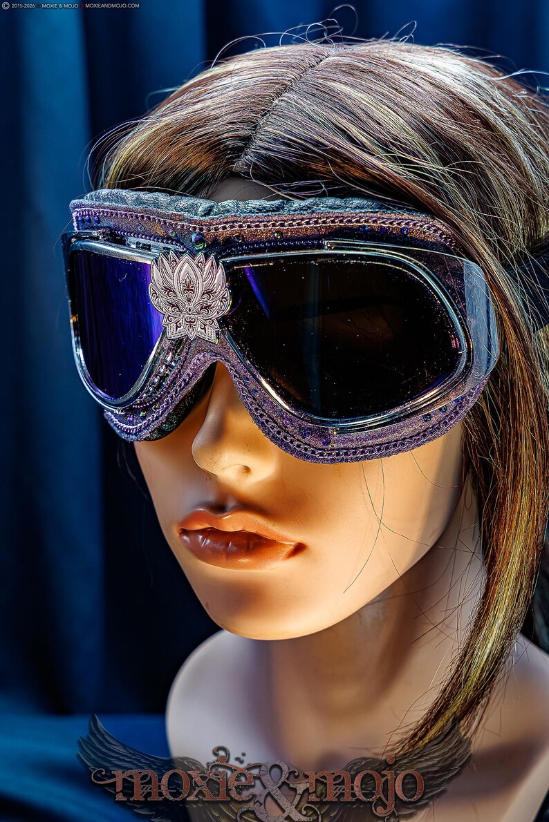 Puede incluir: Gafas protectoras de color morado con lentes oscuras y un emblema plateado en forma de loto centrado en el puente. El marco presenta un borde morado texturizado, detalles met&aacute;licos intrincados y peque&ntilde;os cristales incrustados.