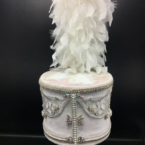 May include: A white drum hat with a white feather plume. The hat is decorated with white embroidery, iridescent rhinestones, and pearl beads.