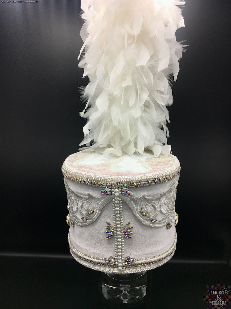 May include: A white drum hat with a white feather plume. The hat is decorated with white embroidery, iridescent rhinestones, and pearl beads.