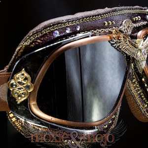 K&ouml;nnte beinhalten: Schutzbrille mit dunkel get&ouml;nten Gl&auml;sern, kupferfarbenem Rahmen und braunem elastischem Band. Die Au&szlig;enseite ist mit goldfarbenen Metallfl&uuml;geln, filigranen Verzierungen, kleinen klaren Kristallen und einem strukturierten dunkelbraunen Rand versehen.