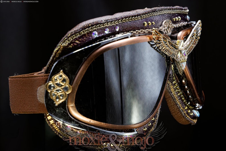 K&ouml;nnte beinhalten: Schutzbrille mit dunkel get&ouml;nten Gl&auml;sern, kupferfarbenem Rahmen und braunem elastischem Band. Die Au&szlig;enseite ist mit goldfarbenen Metallfl&uuml;geln, filigranen Verzierungen, kleinen klaren Kristallen und einem strukturierten dunkelbraunen Rand versehen.
