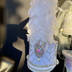May include: A white and silver drum major hat with a large iridescent jewel and white feathers. The hat is decorated with white beads and a white velvet band.