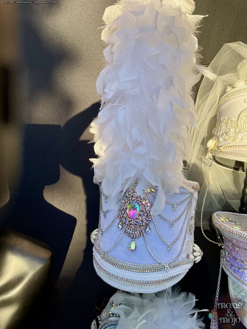 May include: A white and silver drum major hat with a large iridescent jewel and white feathers. The hat is decorated with white beads and a white velvet band.