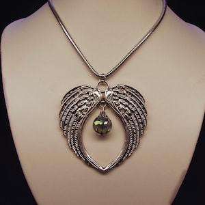 Puede incluir: Un collar de plata con un colgante en forma de coraz&oacute;n con dos alas de &aacute;ngel y una gota de cristal verde.