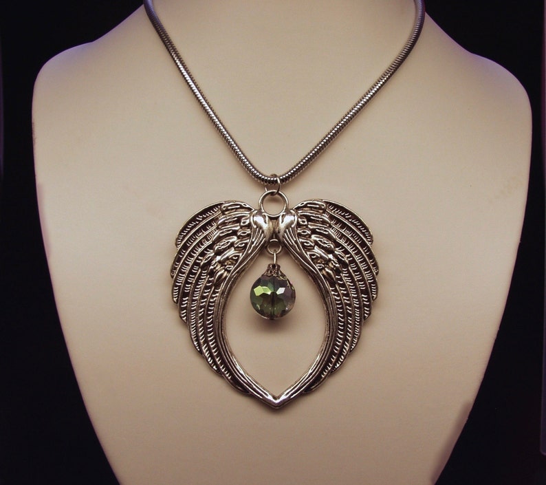 Puede incluir: Un collar de plata con un colgante en forma de coraz&oacute;n con dos alas de &aacute;ngel y una gota de cristal verde.