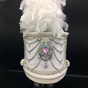 May include: White drum major hat with a white feather plume and a large iridescent rhinestone embellishment. The hat is decorated with white beads and rhinestones.