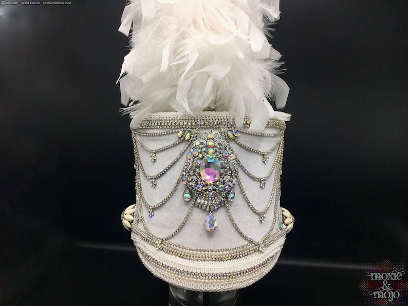 May include: White drum major hat with a white feather plume and a large iridescent rhinestone embellishment. The hat is decorated with white beads and rhinestones.