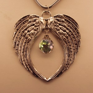 Puede incluir: Un collar de plata con un colgante en forma de coraz&oacute;n que presenta dos alas de &aacute;ngel y un cristal verde en el centro.