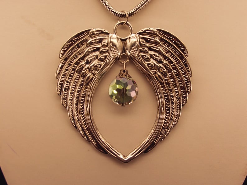 Puede incluir: Un collar de plata con un colgante en forma de coraz&oacute;n que presenta dos alas de &aacute;ngel y un cristal verde en el centro.
