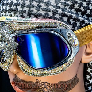 Puede incluir: Gafas protectoras con lentes azul oscuro y una montura decorada con cristales transparentes y pedrer&iacute;a. Incluyen una cinta el&aacute;stica dorada y se llevan sobre un pa&ntilde;uelo con estampado en blanco y negro que muestra el texto moxie & mojo.